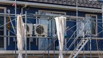 A worker in protective gear is painting the exterior of a building using scaffolding and air conditioning units are visible.