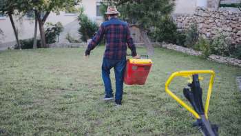 A person in a plaid shirt and hat walks across a lawn, carrying a red and yellow container.