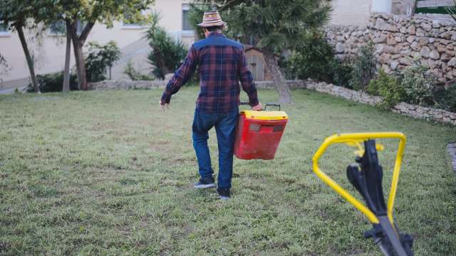 A person in a plaid shirt and hat walks across a lawn, carrying a red and yellow container.