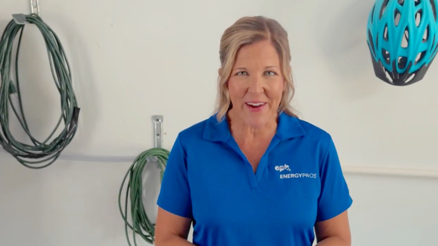 A smiling woman in a blue polo shirt stands in front of a wall with various cords and a bike helmet.