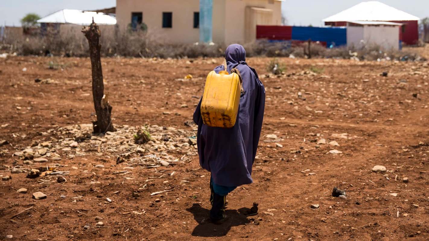 A person dressed in a dark cloak walks across a dry, rocky landscape, carrying a yellow water container.