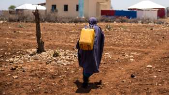 A person dressed in a dark cloak walks across a dry, rocky landscape, carrying a yellow water container.