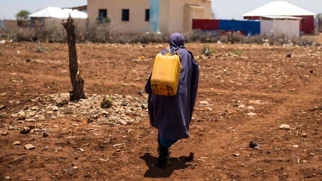 A person dressed in a dark cloak walks across a dry, rocky landscape, carrying a yellow water container.