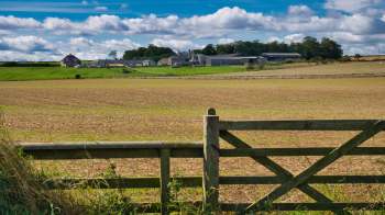 A wide view of a rural landscape featuring a farm, fields, and a wooden gate against a cloudy sky.