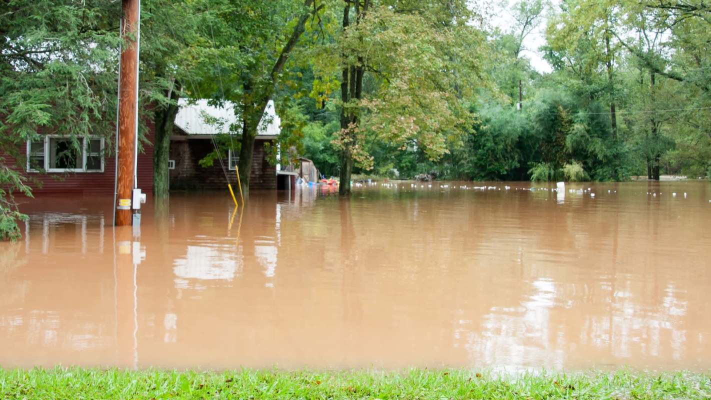 A house partially submerged in muddy floodwaters, surrounded by trees and debris.