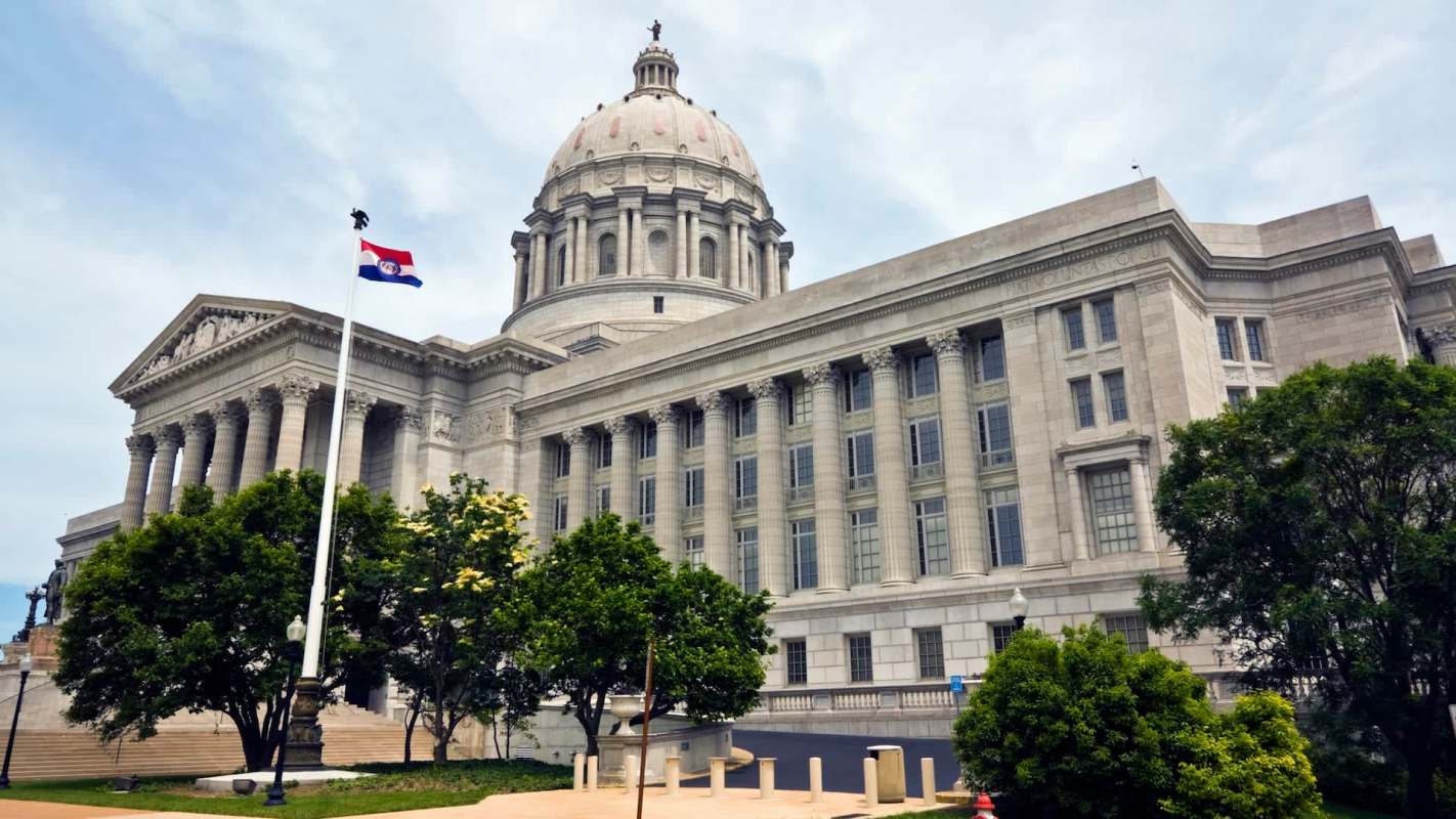 The Missouri State Capitol building features a large dome and classical architectural details, surrounded by green trees.