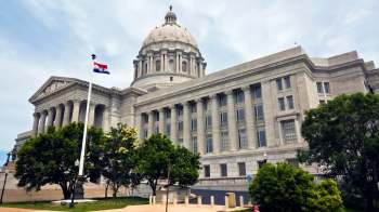 The Missouri State Capitol building features a large dome and classical architectural details, surrounded by green trees.