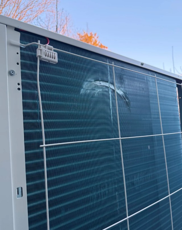 A close-up of an HVAC unit with fins  bent out of place, in front of a clear blue sky. 