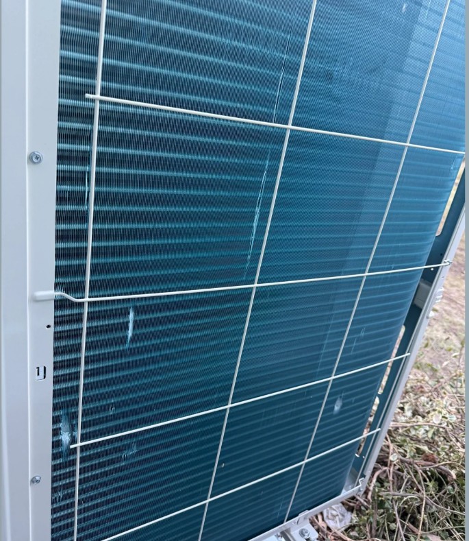 An HVAC unit with fins bent out of place, situated on brown vegetation. 