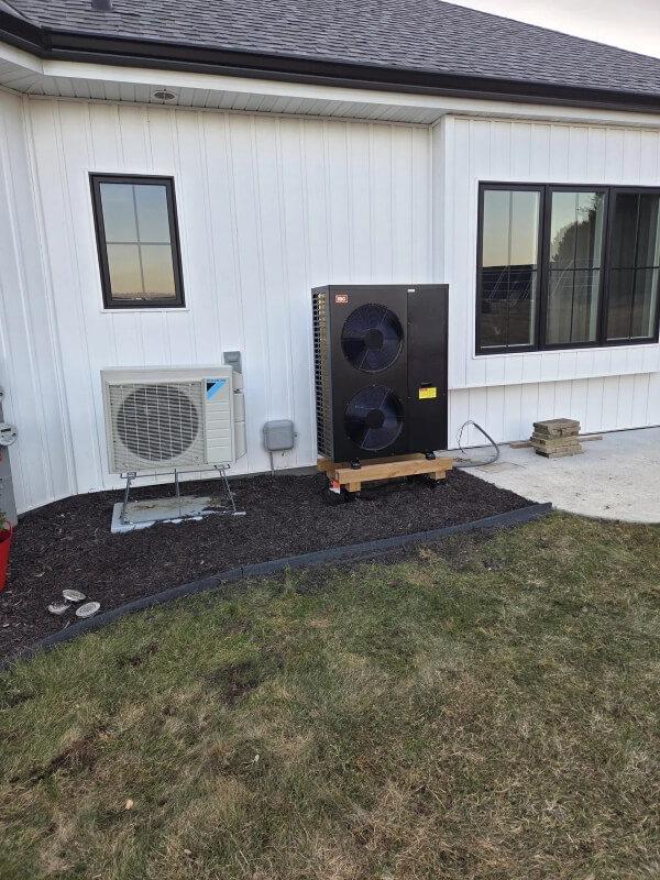 A black hydronic heat pump in front of a white house with black window frames. 