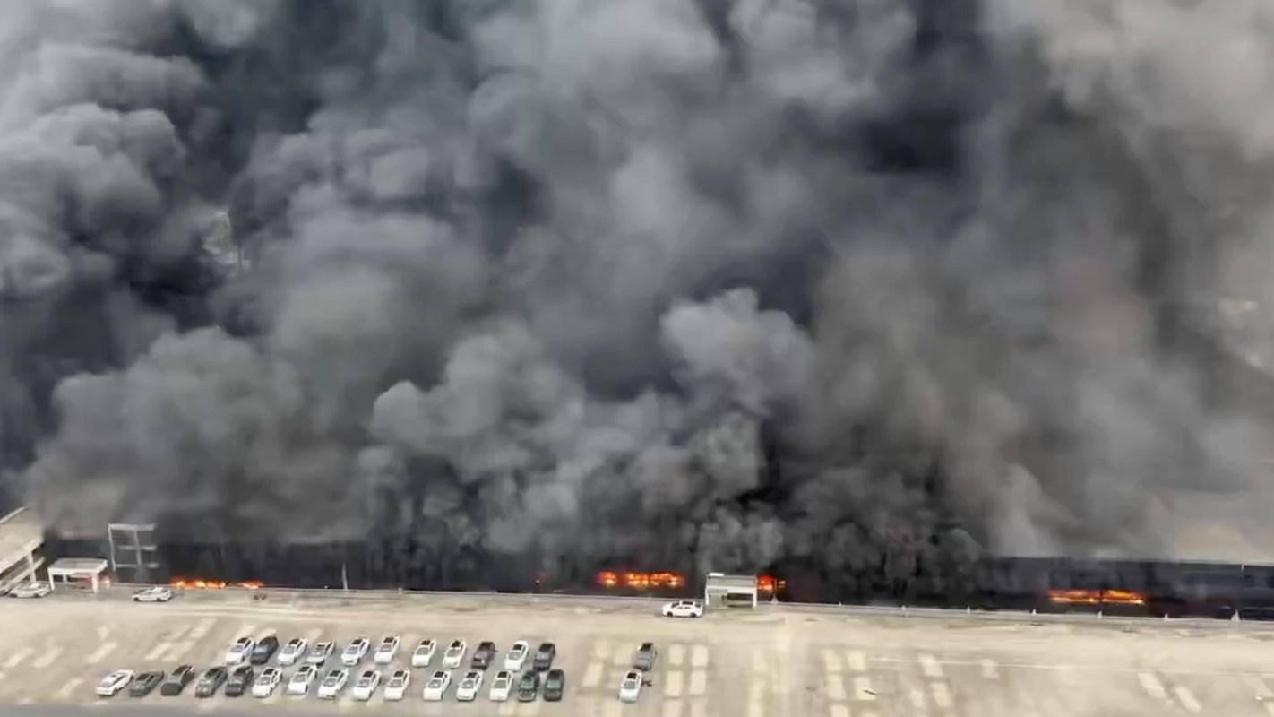 Thick black smoke billows over a parking lot, with visible flames near a structure.