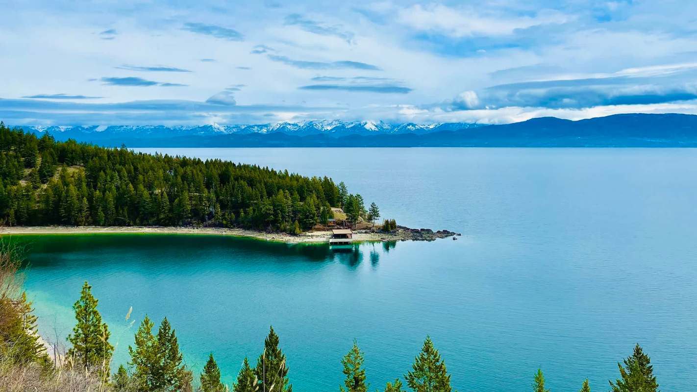 A serene Flathead Lake in Montana surrounded by evergreen trees and distant snow-capped mountains under a partly cloudy sky.
