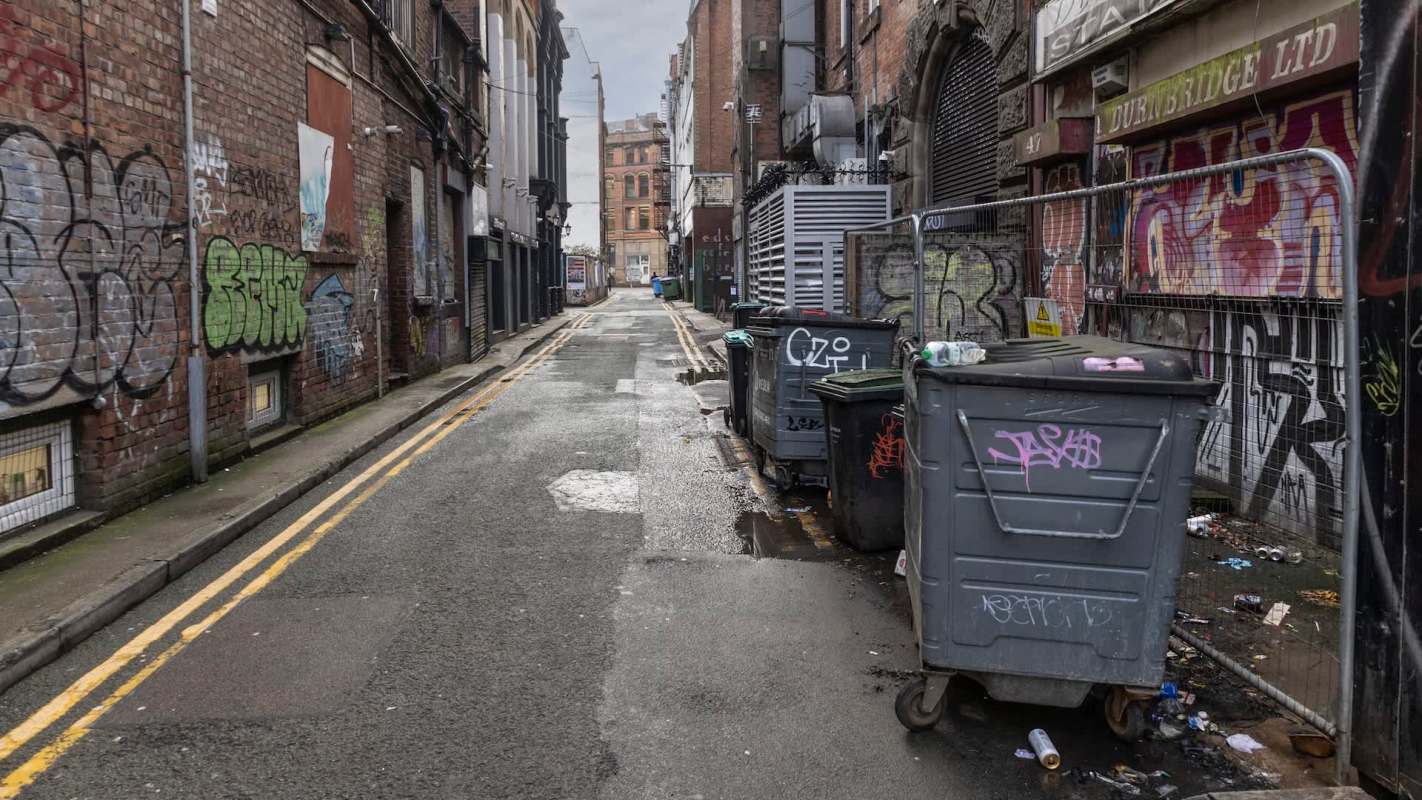 A narrow urban alley lined with graffiti and trash bins, flanked by brick buildings under a cloudy sky.