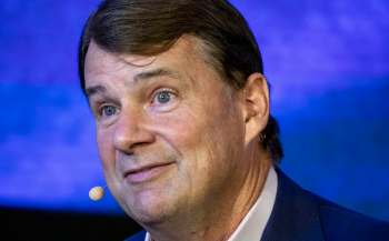 Ford CEO Jim Farley, with short, dark hair and a slight smile, speaks into a microphone against a blue backdrop.