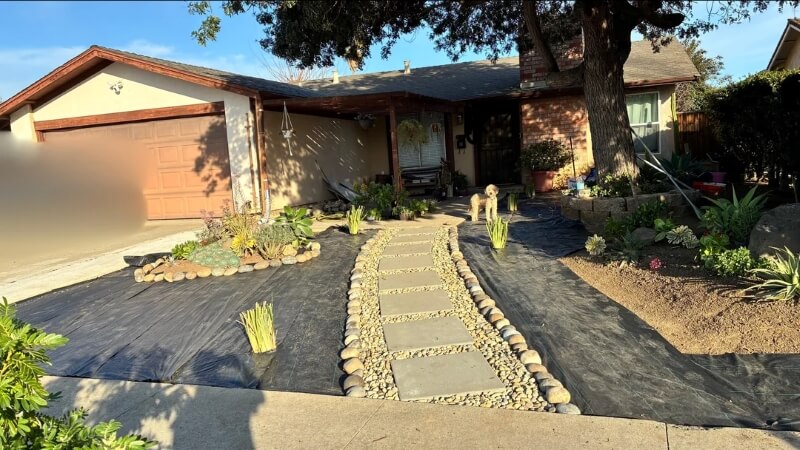 A front yard featuring a stone path and drought-tolerant plants, covered mostly in black fabric. 
