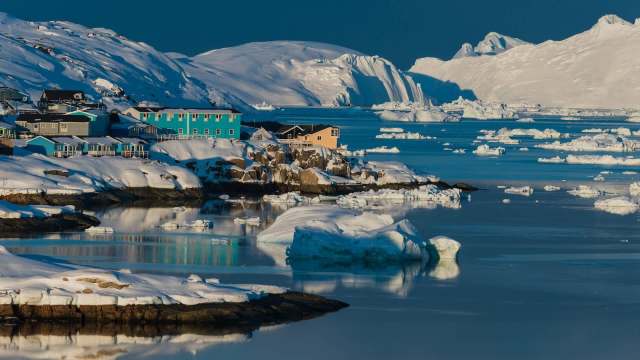 Colorful houses on a snowy shoreline in Greenland reflect in calm waters, surrounded by icebergs and distant mountains.
