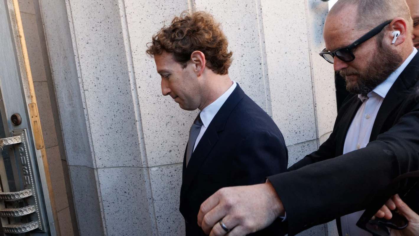 Mark Zuckerberg in a suit walks past security with his head down, accompanied by another man adjusting his glasses.
