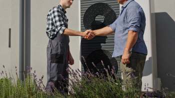 Two men shake hands in a garden, surrounded by lavender, with a heat pump in the background.