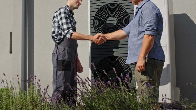 Two men shake hands in a garden, surrounded by lavender, with a heat pump in the background.