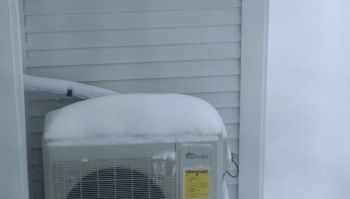 An air conditioning unit covered in snow next to a house's siding on a winter day.