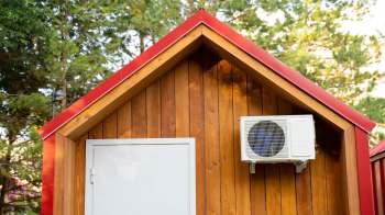 A wooden cabin with a red roof and a heat pump unit mounted outside.