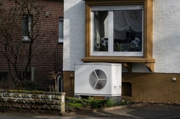 An air conditioning unit installed outside a house next to a window with curtains and visible plants inside.