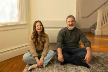 A woman and a man, Mary-Ann Rau and Brad Hall, sit on the floor, smiling together in a warmly lit room.