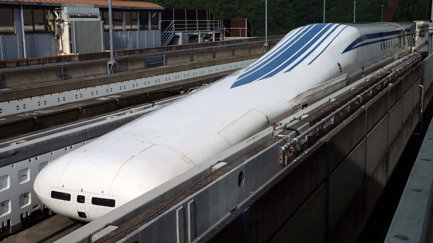 A sleek maglev train displayed on tracks within a maintenance facility surrounded by greenery.