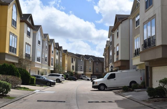 A residential street lined with multi-colored townhouses and parked vehicles under a partly cloudy sky.