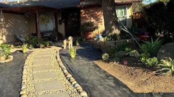 A front yard featuring a stone path, potted plants, and a small dog standing near the entrance.