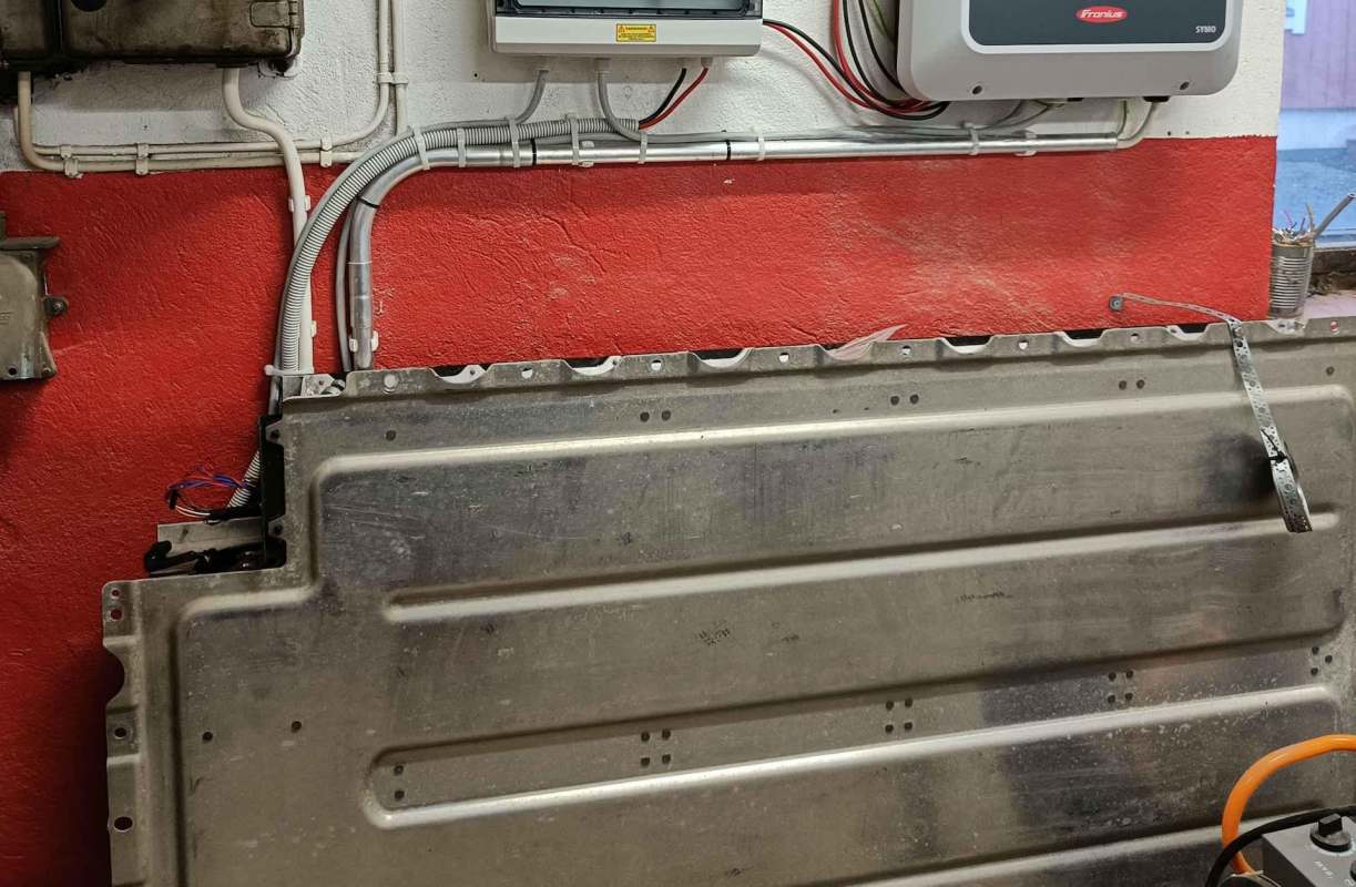 A metallic panel on a red wall, surrounded by equipment including a used EV battery.