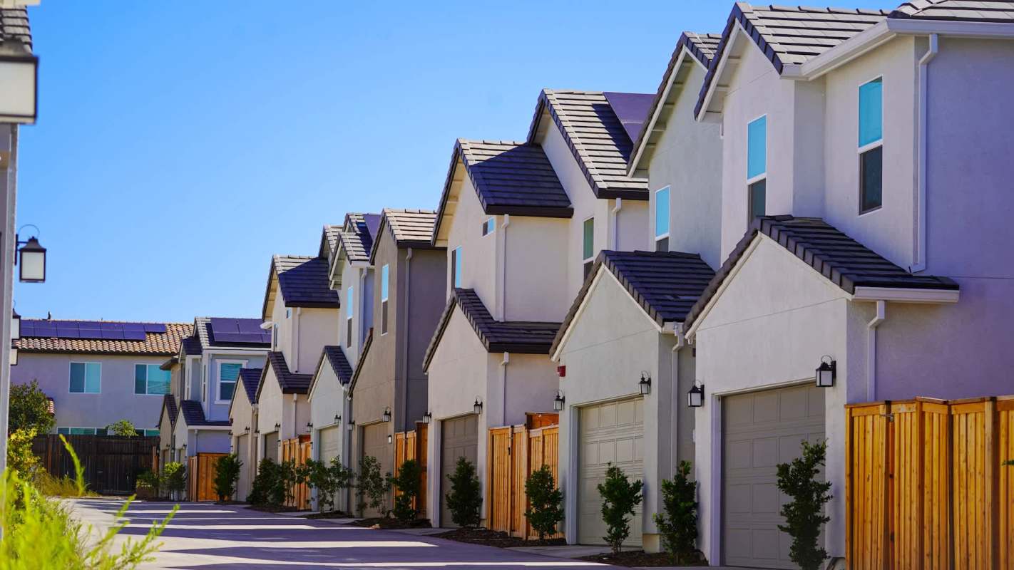 A row of modern townhouses with pitched roofs and garages lining a sunny street.