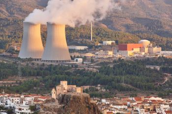A nuclear power plant with cooling towers emits steam, set against a backdrop of hills and a nearby village.