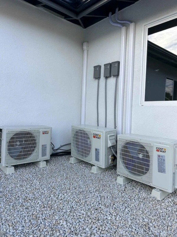 Three mini-split heat pumps sitting on gravel in front of two white walls. 
