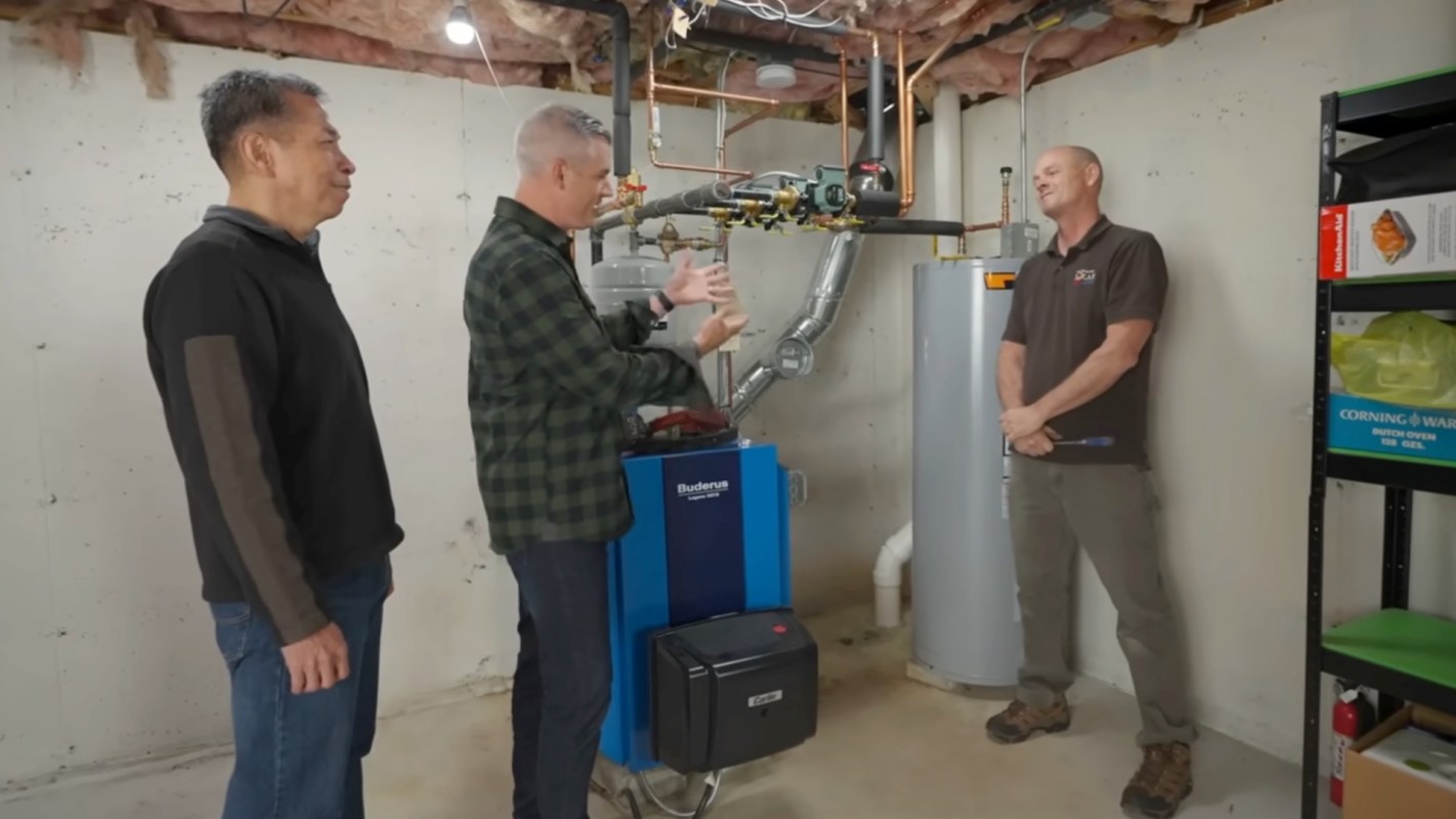 Three men converse in a basement with a heating system, discussing its features and functionality.