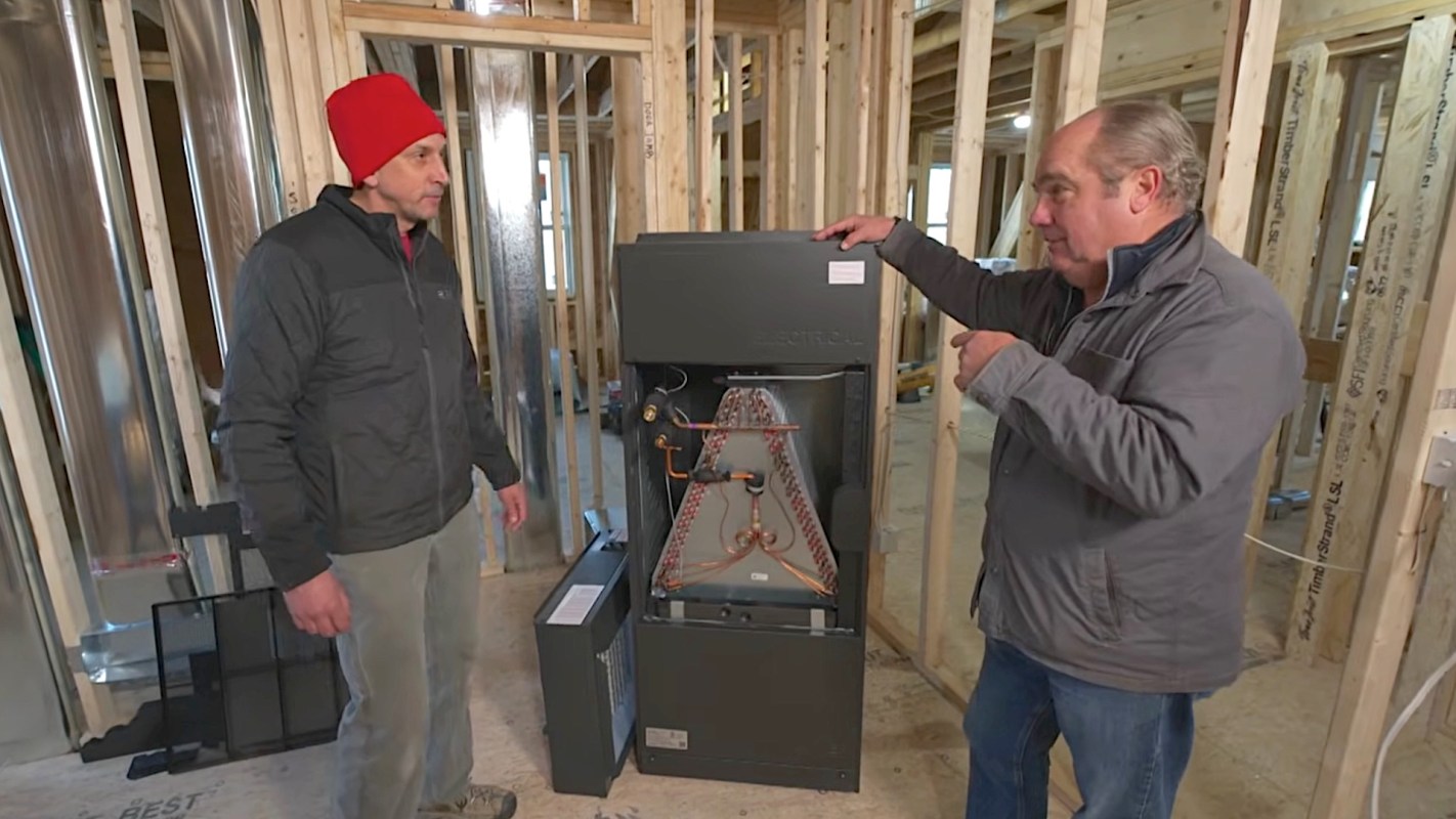Two men discuss a heating unit inside a partially constructed room with exposed wooden framing.