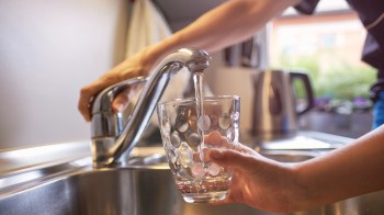 A person fills a clear glass with water from a kitchen faucet.