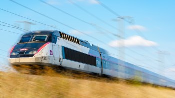 A speeding train passes through a grassy landscape under a blue sky with scattered clouds.