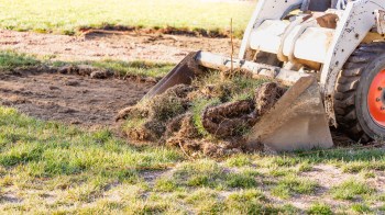 A skid steer loader lifting a clump of sod and soil in a grassy area.