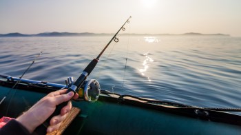 A person holding a fishing rod over calm water with sun reflections and distant hills.