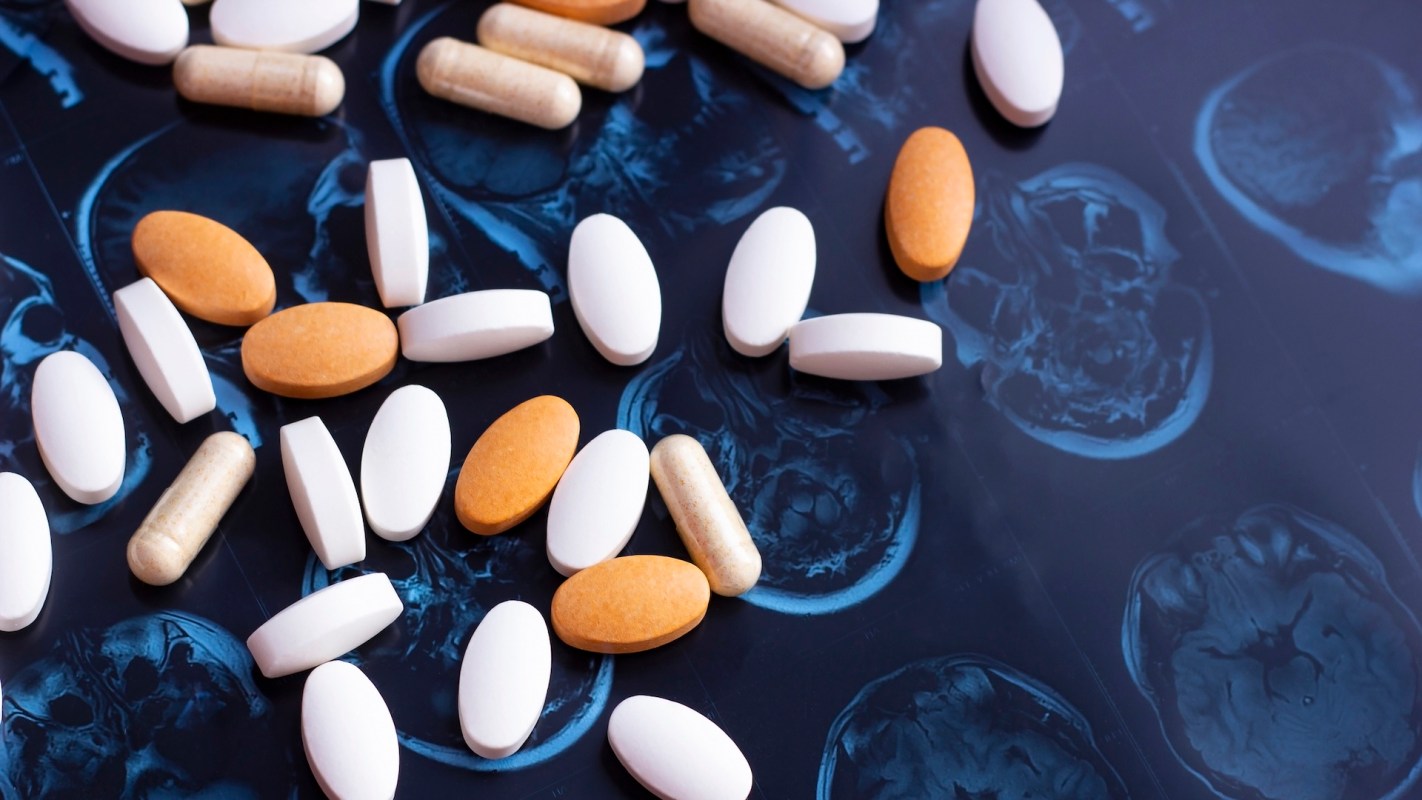 Assorted pills and capsules scattered over a dark surface featuring MRI brain scans.