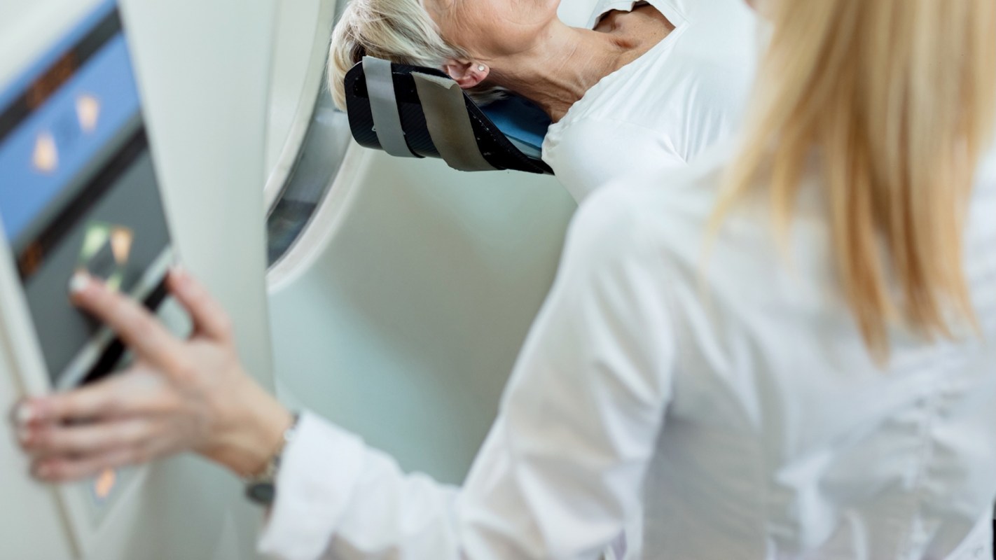 A patient reclines in a medical bed for a CT scan while a radiologist adjusts controls nearby.