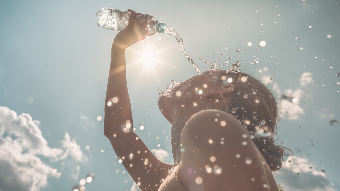 A woman pours water over her head, surrounded by sparkling droplets and sunlight against a bright blue sky.