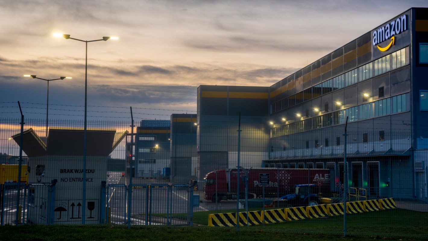 An Amazon distribution center at dusk, featuring security fencing and illuminated loading areas.