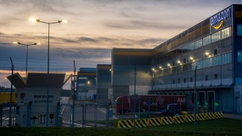 An Amazon distribution center at dusk, featuring security fencing and illuminated loading areas.