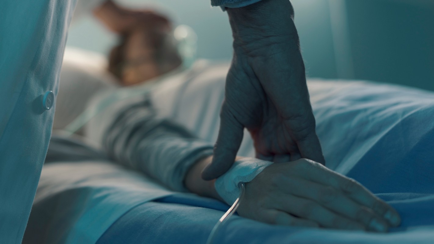 A hospitalized patient lying in a hospital bed, with a doctor placing their hands on his forehead and wrist.