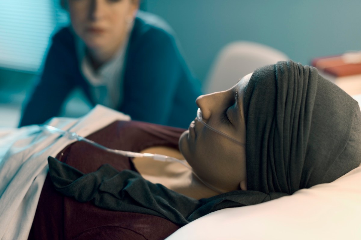 A female cancer patient in a hospital bed, resting with an oxygen tube, while a concerned figure watches in the background.