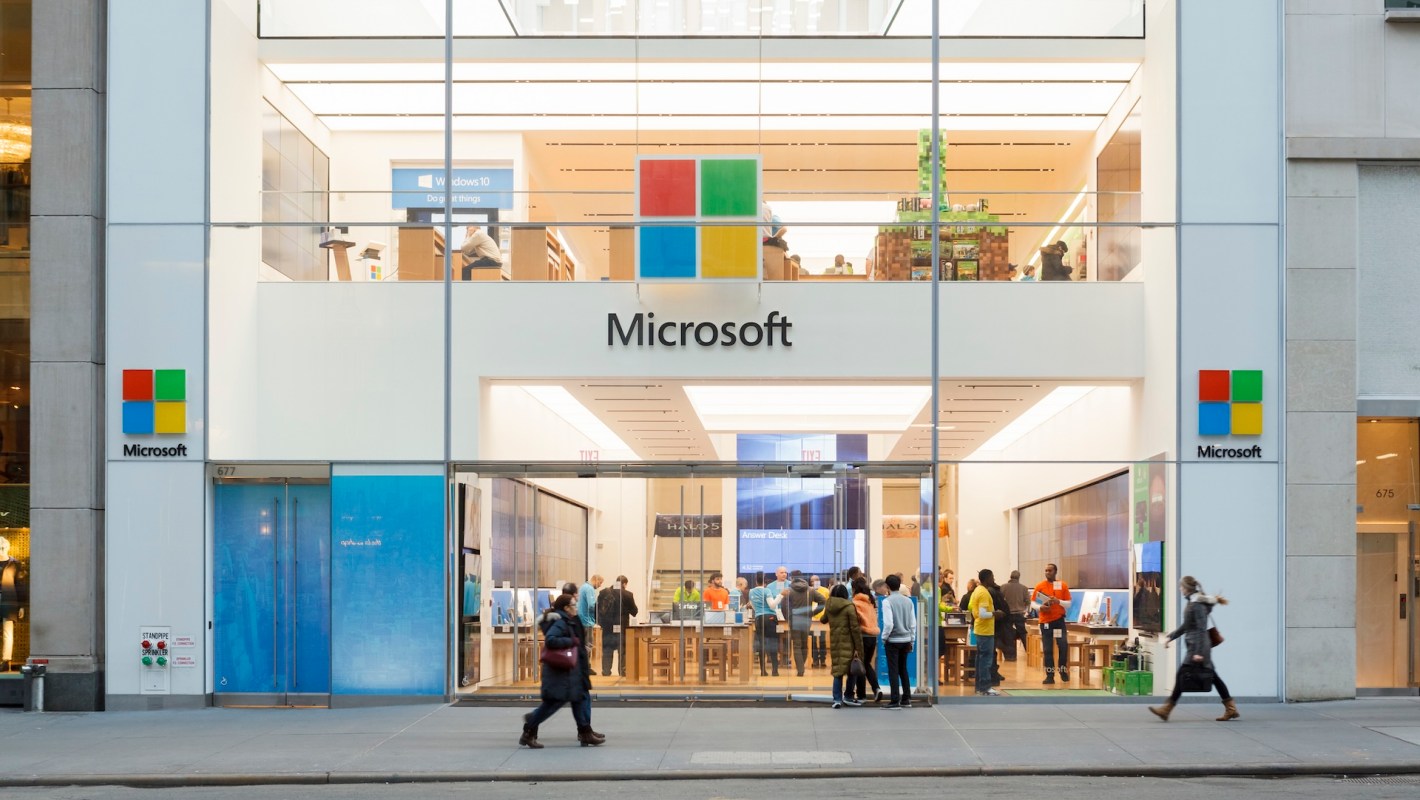 A modern Microsoft store exterior with people inside and outside, showcasing products and a bright design.