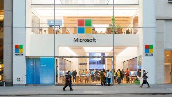 A modern Microsoft store exterior with people inside and outside, showcasing products and a bright design.