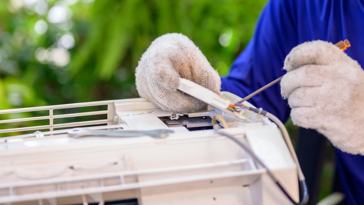 A technician in gloves repairs an air conditioning unit with tools in a green outdoor setting.